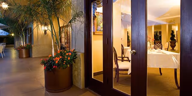 View of an outdoor patio area with large potted plants and flowers next to glass doors leading into a warmly lit dining room with a table set for a meal and chairs around it.