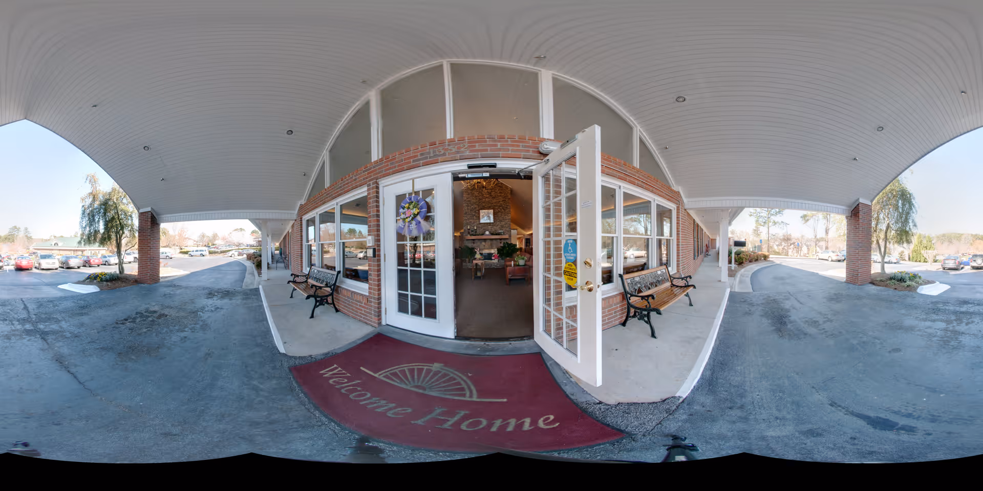 Entrance to a senior living facility with a covered driveway and a red welcome mat that says 'Welcome Home'. The entrance has double glass doors, one of which is open, and two benches on either side. Inside, a cozy lobby area with a stone fireplace and seating is visible. The exterior walls are brick, and there is a parking lot with cars and trees in the background.