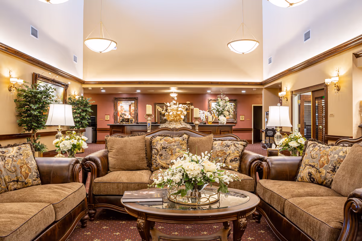 A spacious and elegantly decorated living room area with brown leather and fabric sofas arranged around a glass coffee table adorned with a floral centerpiece. The room features warm lighting from ceiling fixtures and table lamps, with framed artwork and plants adding to the cozy ambiance.
