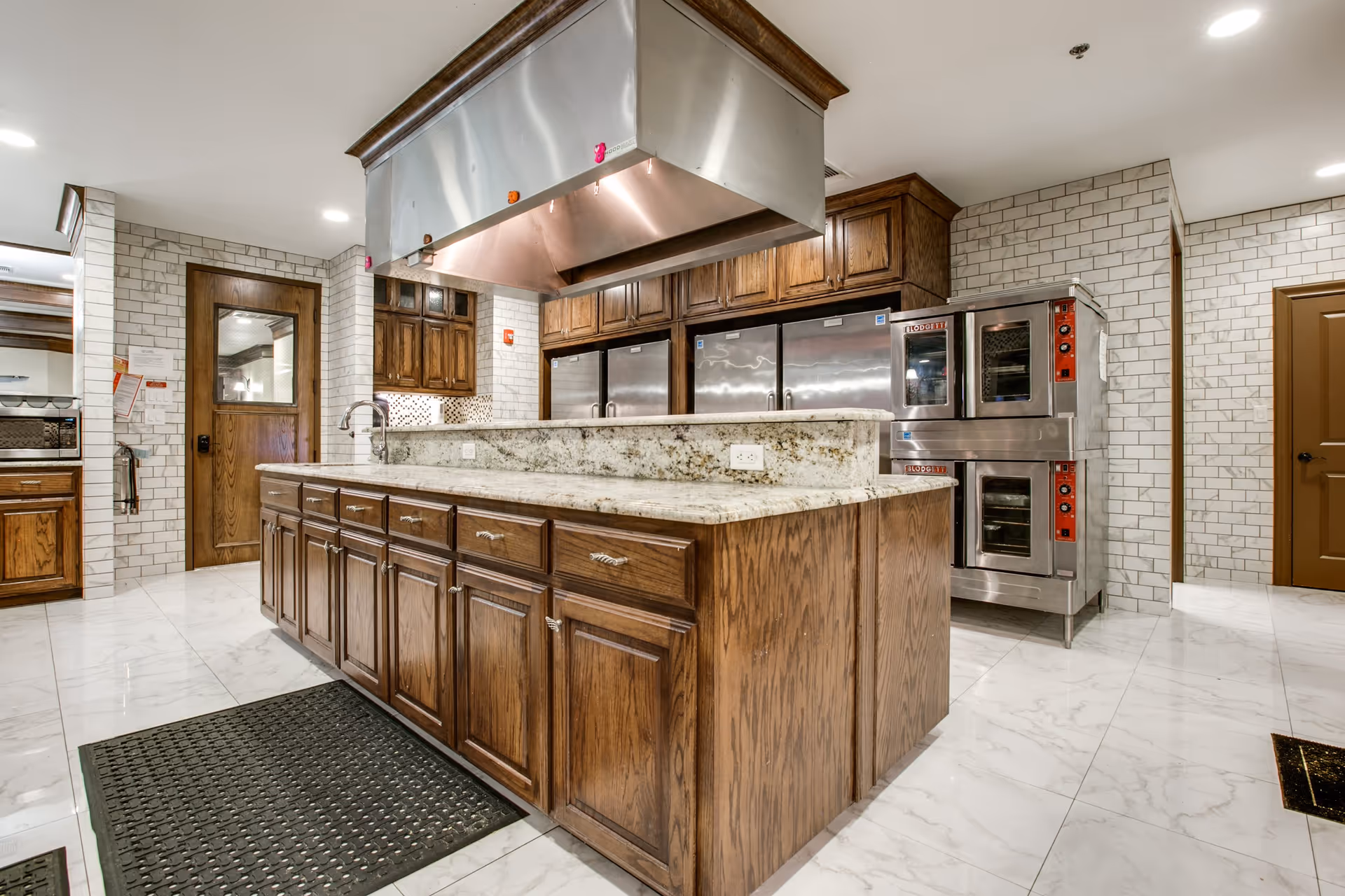 A spacious kitchen with wooden cabinets, a large granite countertop island, stainless steel appliances including refrigerators and ovens, white tiled walls, and a white tiled floor with a black rubber mat in front of the island.