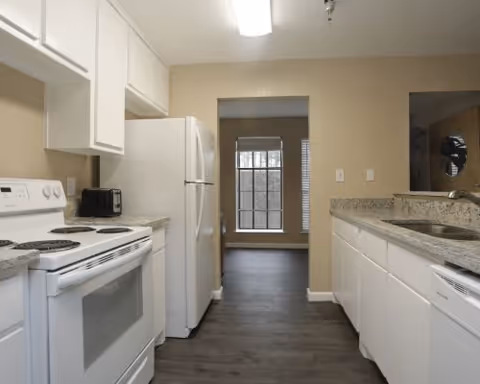 A clean and modern kitchen with white cabinets, a white electric stove, a white refrigerator, a microwave, granite countertops, and a stainless steel sink. The kitchen has wood flooring and beige walls, with a window visible in the adjacent room.