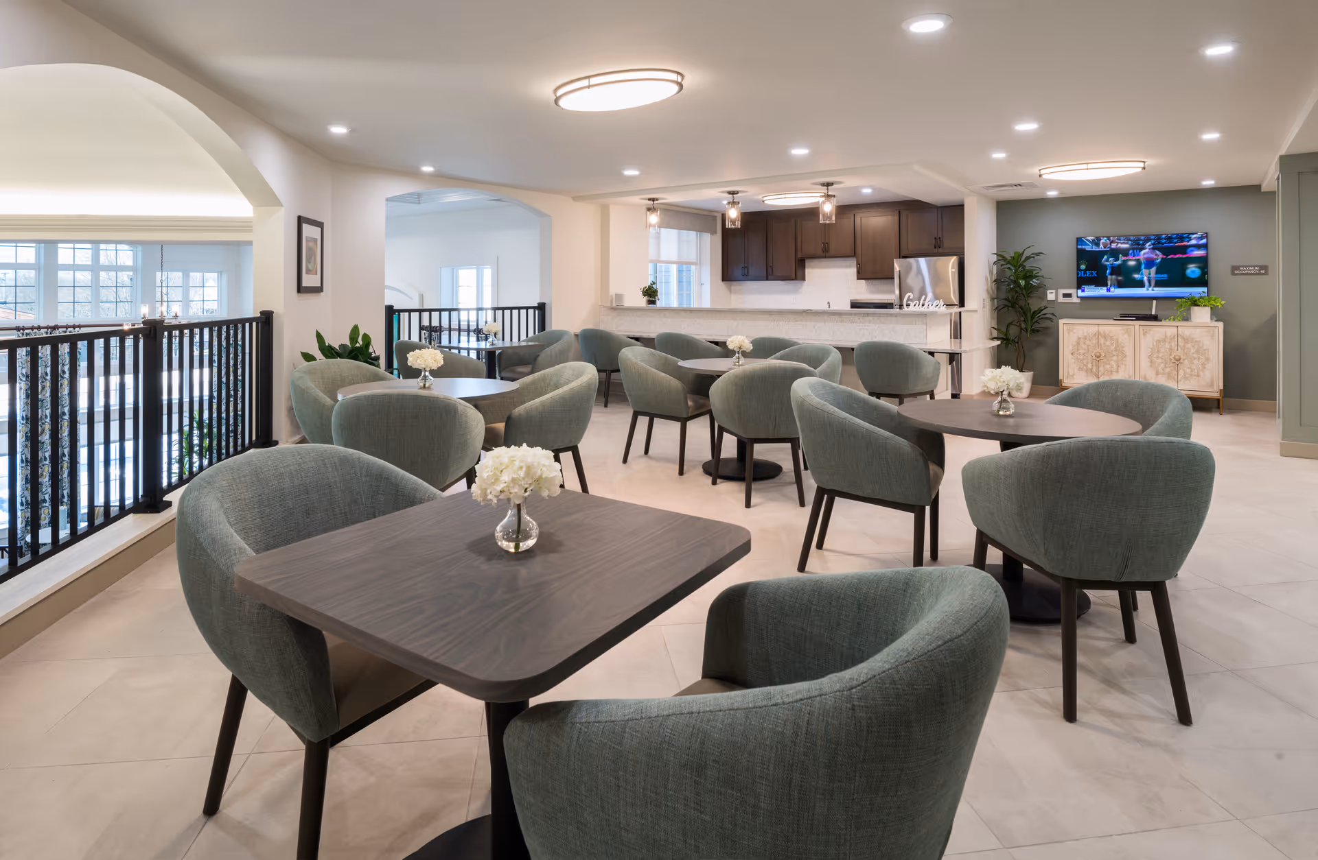 A modern senior living facility common area with multiple round and square tables surrounded by green upholstered chairs. Each table has a small vase with white flowers. The space features a kitchen area with dark wood cabinets and stainless steel appliances in the background. A flat-screen TV is mounted on the wall above a decorative cabinet, and there are plants placed around the room. The area is well-lit with ceiling lights and has large windows allowing natural light.