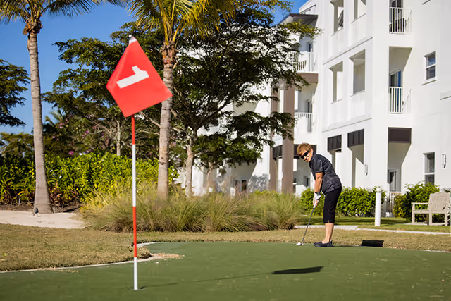 A senior woman playing golf on a putting green near a white multi-story building with balconies. The green has a red flag with the number 1 on it, and there are palm trees and bushes in the background under a clear blue sky.