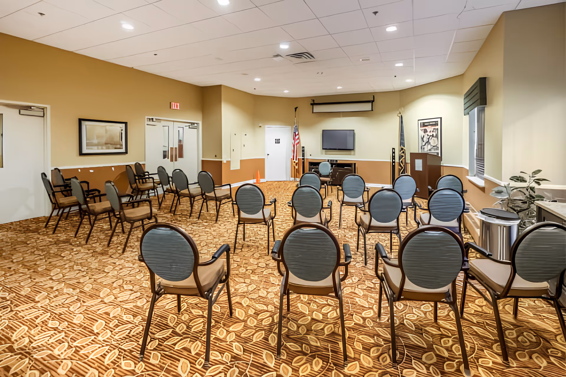 A meeting or activity room with multiple chairs arranged in rows facing a wall-mounted TV and a podium. The room has patterned carpet, beige walls, and ceiling lights. There are two flags near the TV and a window with a valance on the right side.