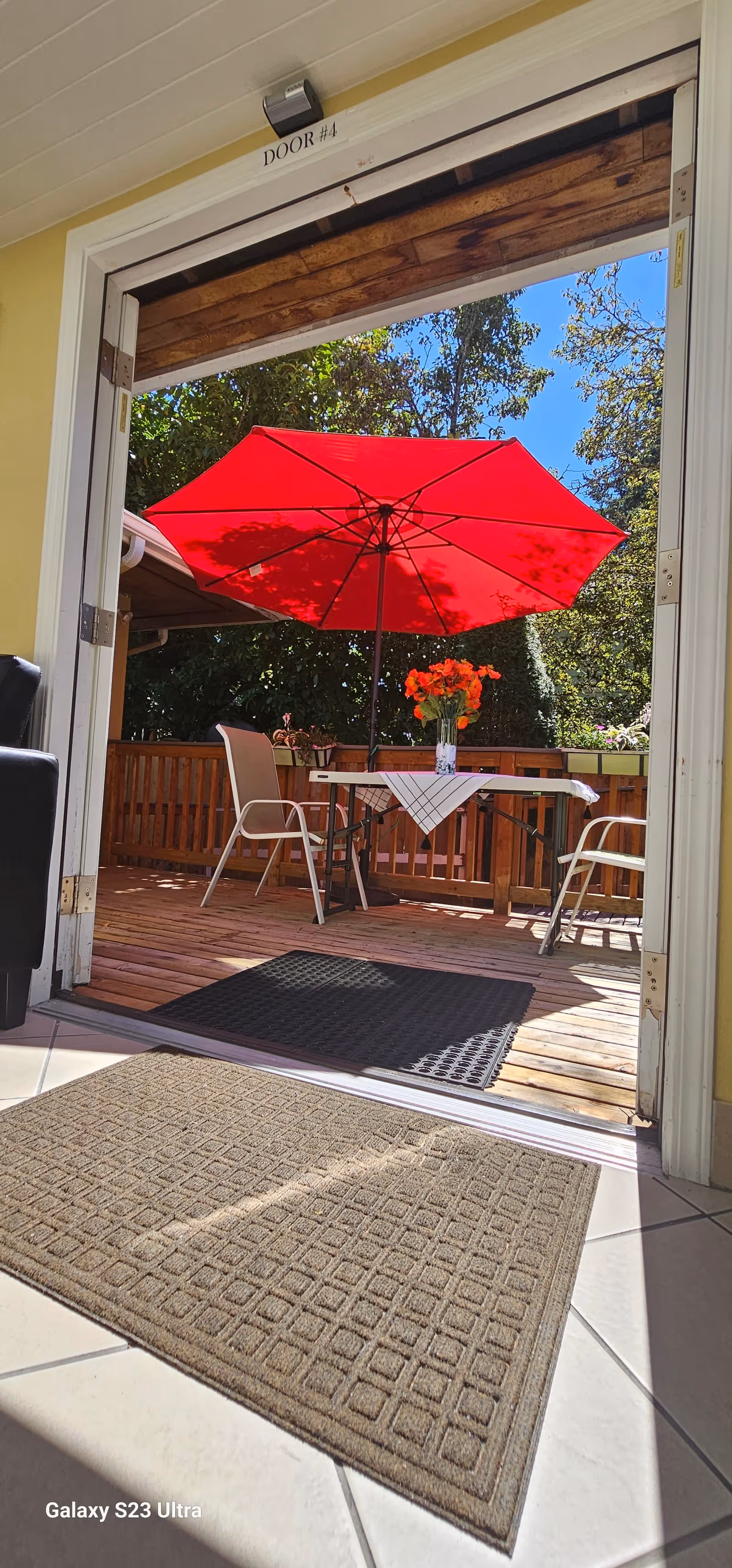 View through an open doorway labeled 'DOOR #4' leading to a wooden deck with a red patio umbrella, a table covered with a white cloth and a vase of orange flowers, and two chairs. The deck is surrounded by greenery and trees under a clear blue sky.