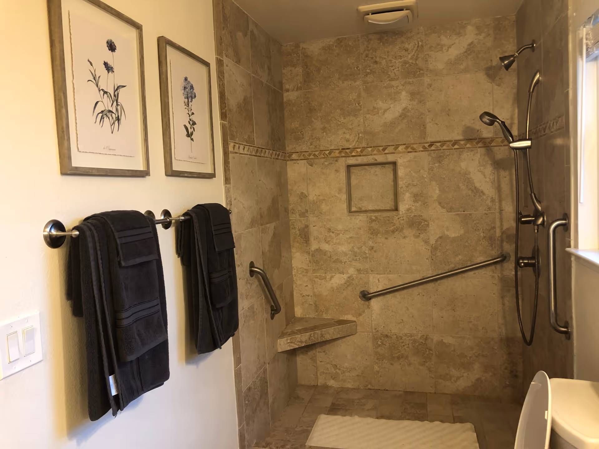 A bathroom with beige tiled walls and floor, featuring a walk-in shower with a built-in corner seat, grab bars, and a handheld showerhead. Two dark towels hang on a towel rack on the left wall beneath two framed botanical prints. A white bath mat is on the floor, and a window is visible on the right side.