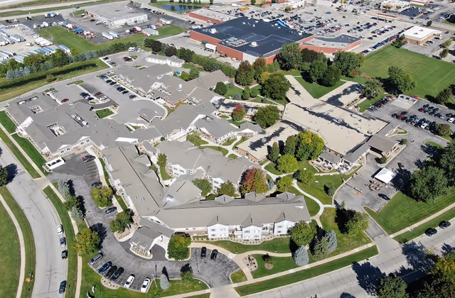 Aerial view of a senior living campus with multiple connected buildings, parking lots, walkways, and surrounding trees and lawns.