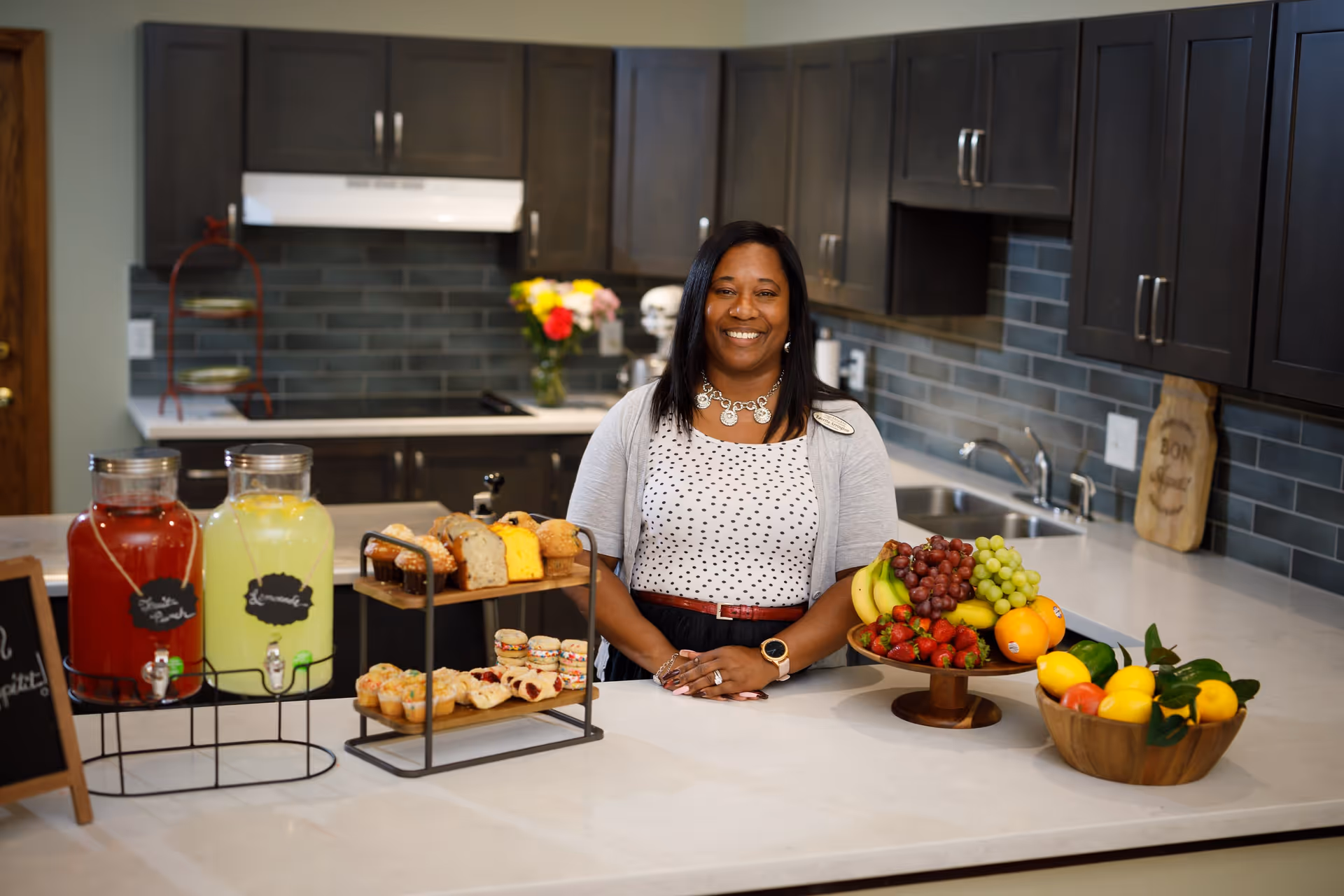 A smiling woman stands behind a kitchen island topped with drinks, pastries, and bowls of fruit in a modern kitchen.