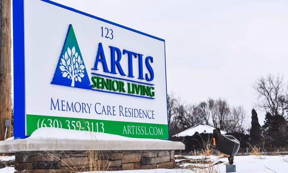 Outdoor sign for Artis Senior Living Memory Care Residence with contact number and website, surrounded by snow and bare trees in the background.