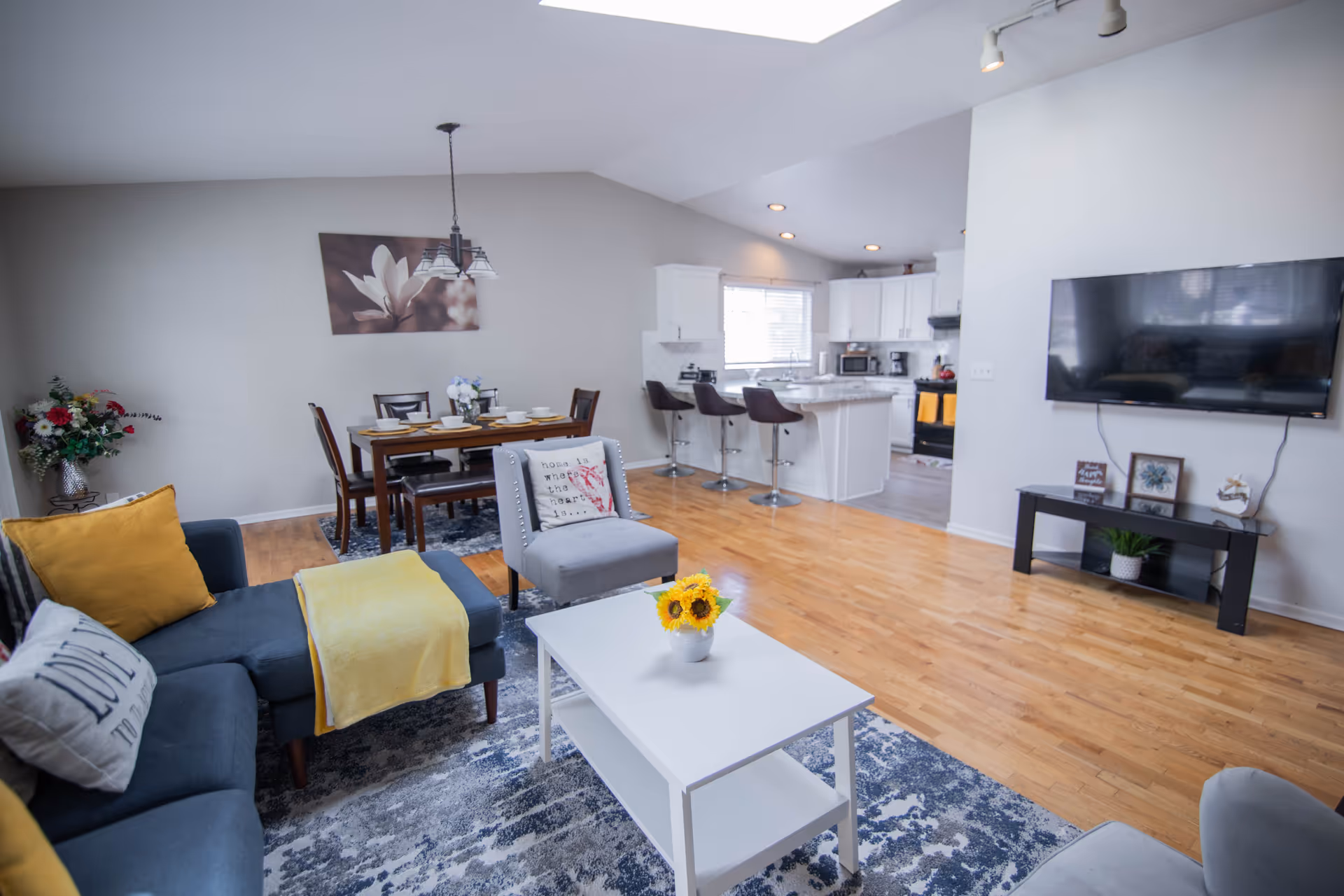 Bright and spacious open living area featuring a blue sectional sofa with yellow and white pillows, a white coffee table with a small sunflower arrangement, a gray armchair, a dining table set with six chairs and tableware, and a kitchen with white cabinets, a breakfast bar with three stools, and modern appliances. A flat-screen TV is mounted on the wall above a black TV stand with decorative items.