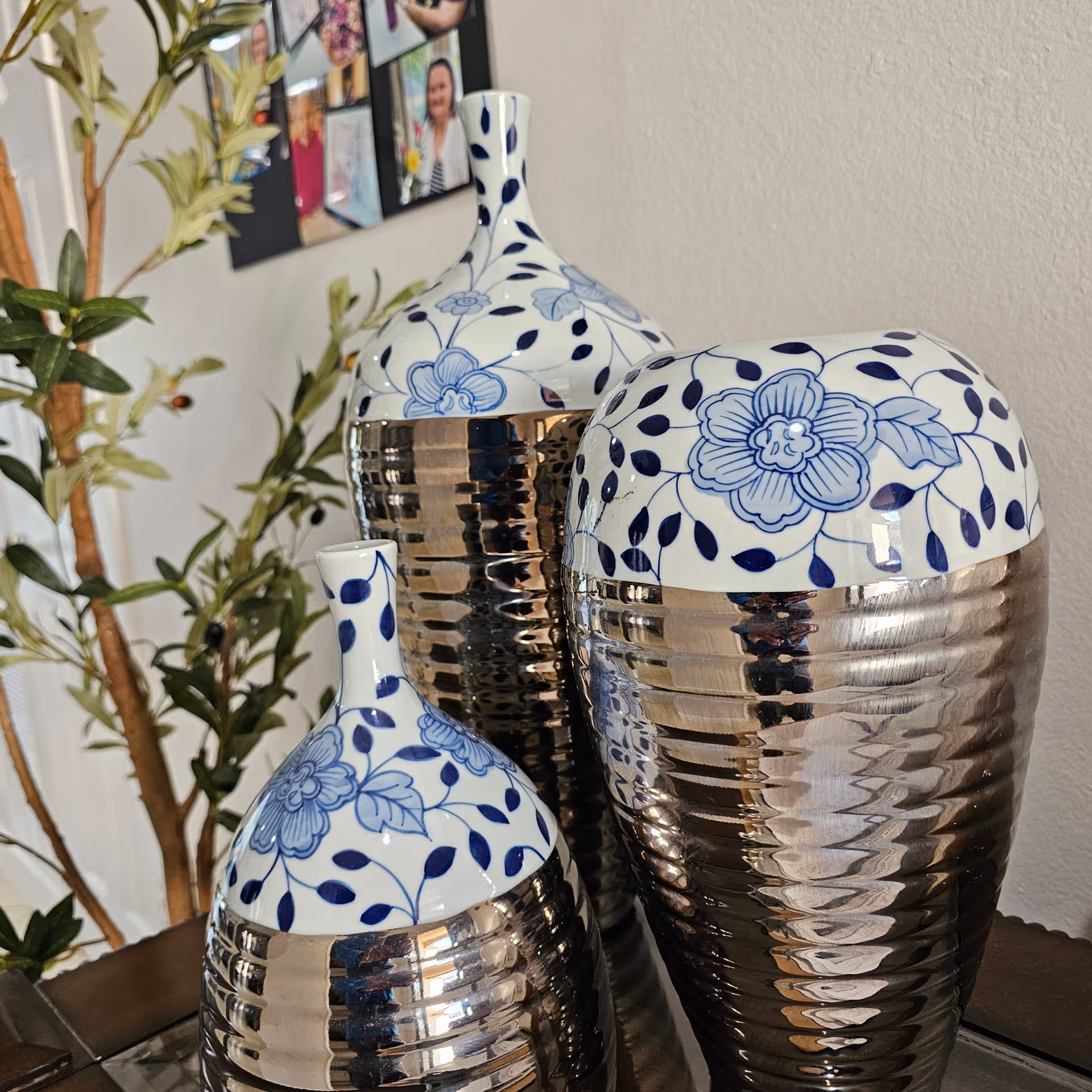 Three decorative vases with blue floral tops and metallic ribbed bases arranged on a table, with a leafy plant and a photo collage on the wall behind them.