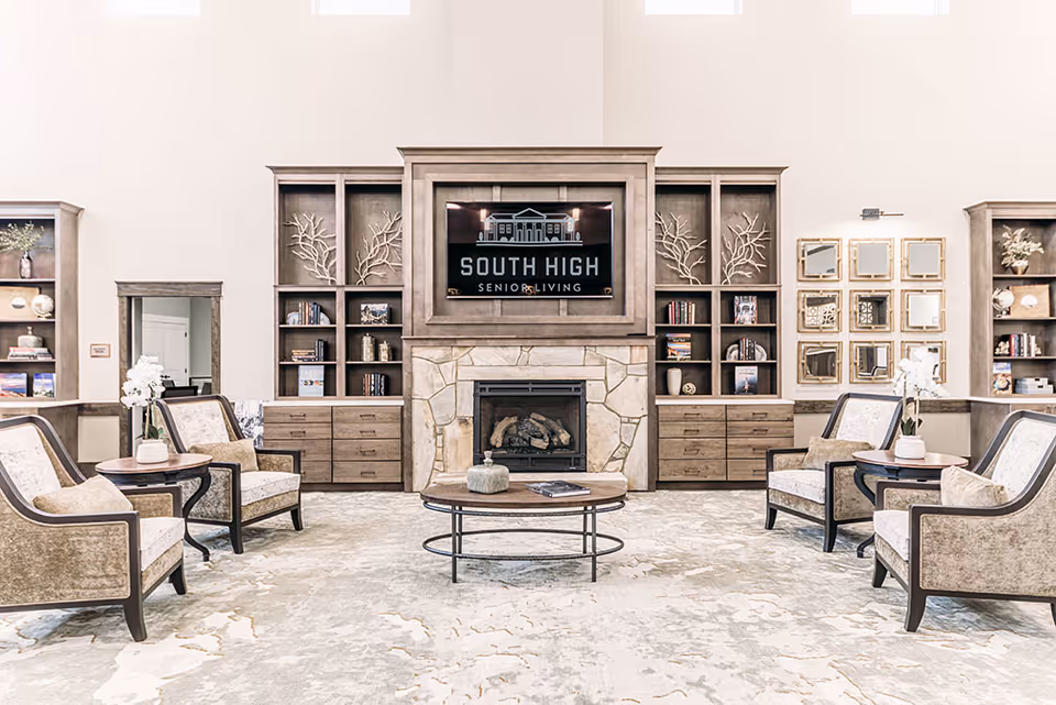 A bright and spacious senior living facility common area with four upholstered armchairs arranged around a round wooden coffee table. The room features built-in wooden shelves with decorative items and books, a stone fireplace, and a large TV above the fireplace displaying the South High Senior Living logo. The walls are light-colored, and there are decorative mirrors on one side.