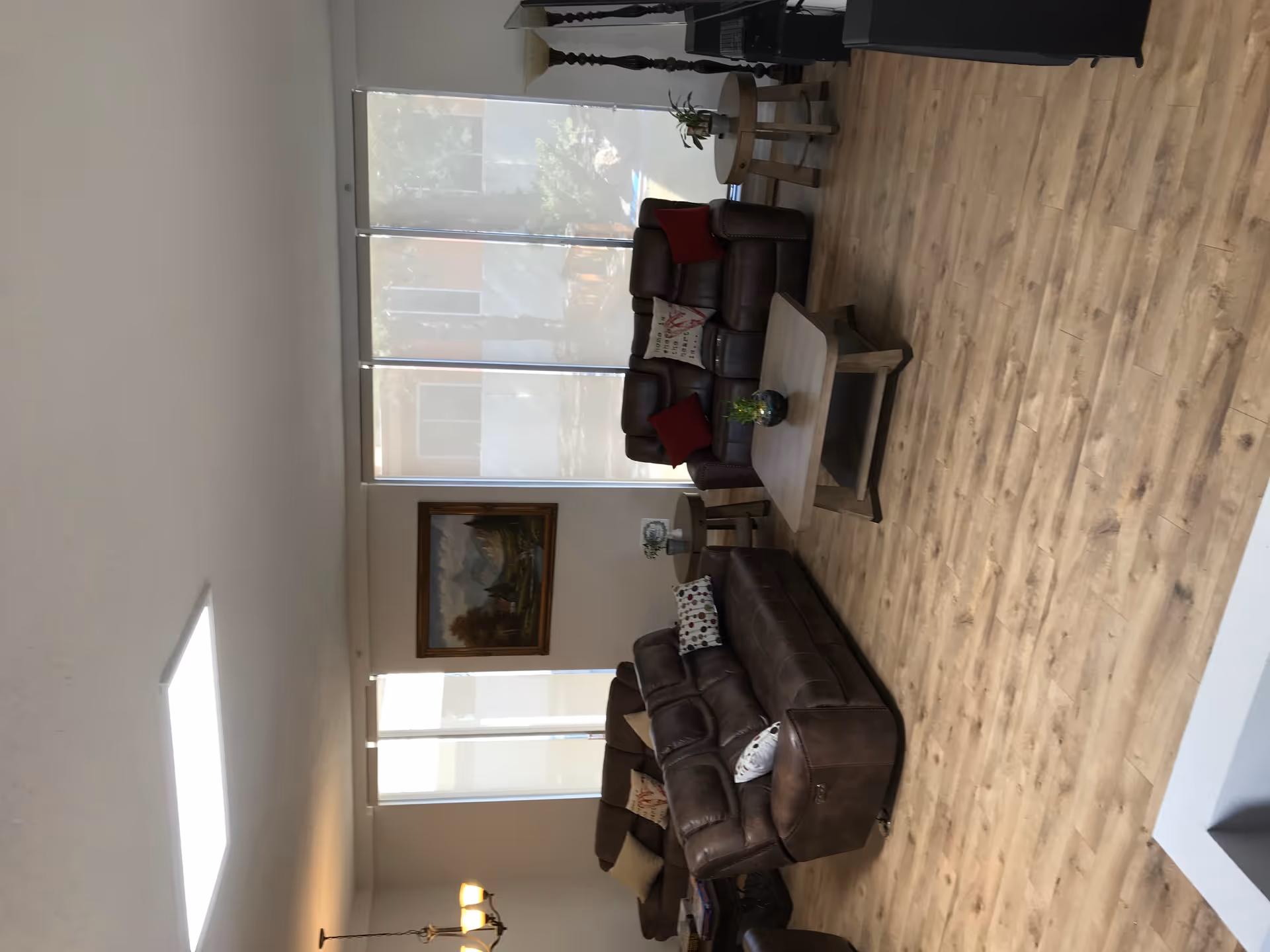 A spacious living room with wooden flooring, two brown leather sofas with decorative pillows, a wooden coffee table with a small plant, two side tables with plants and decor, a floor lamp, large windows with blinds, and a landscape painting on the wall.