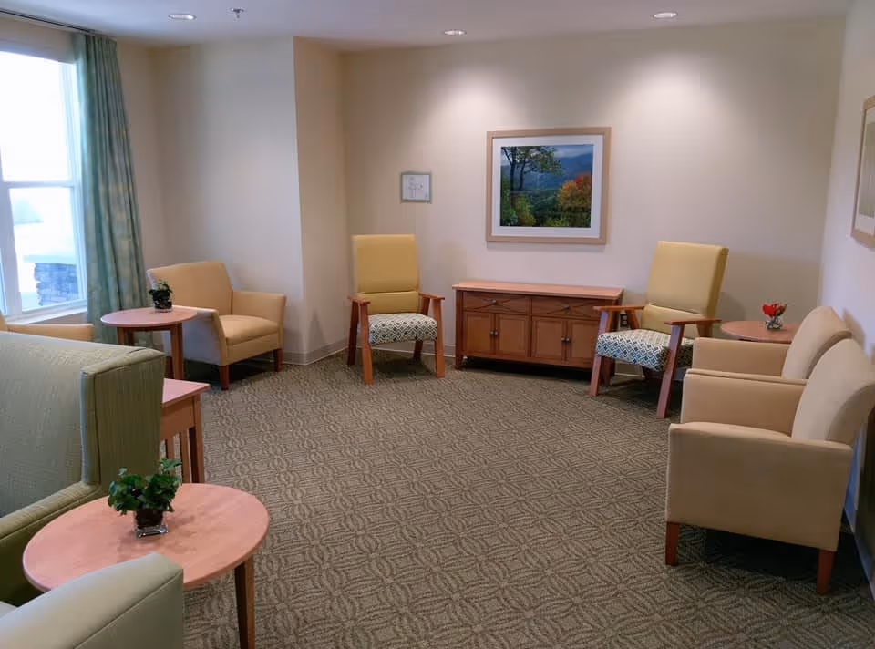A cozy sitting area with multiple armchairs and small round wooden tables, some with small potted plants. The room has beige walls, a patterned carpet, and a framed landscape picture on the wall. A window with green curtains allows natural light to enter the space.