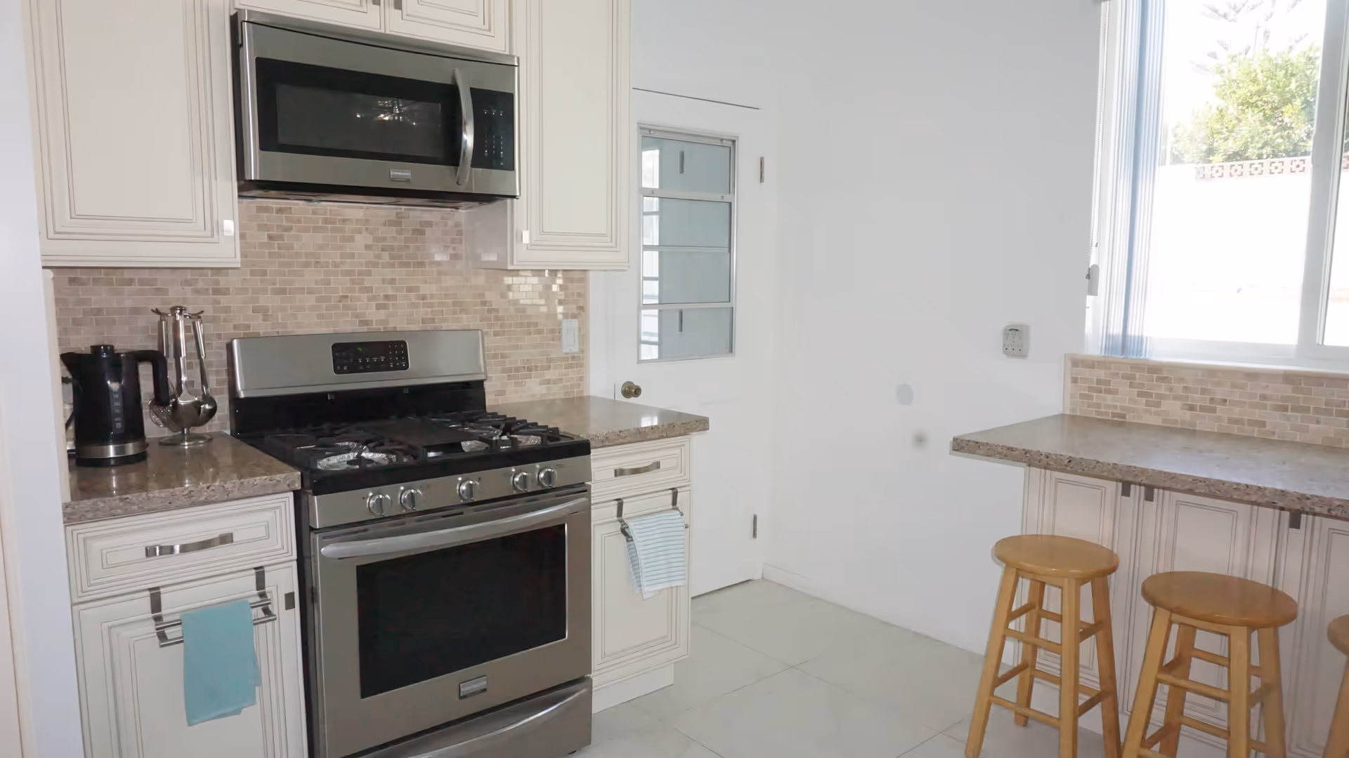 A modern kitchen with stainless steel appliances including a microwave and gas stove. The kitchen features beige tiled backsplash, light-colored cabinetry, and granite countertops. There is a small breakfast bar with two wooden stools and a window letting in natural light.