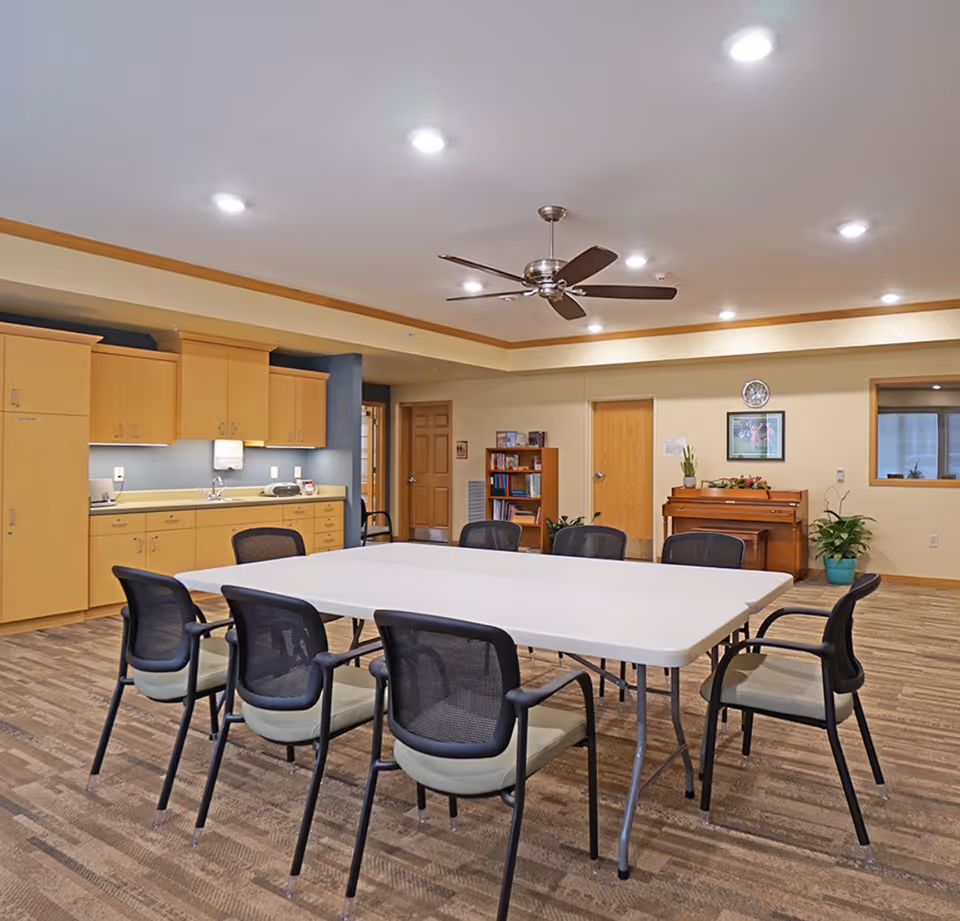A bright communal activity room with folding tables and chairs, a kitchenette, bookshelf, and a piano.