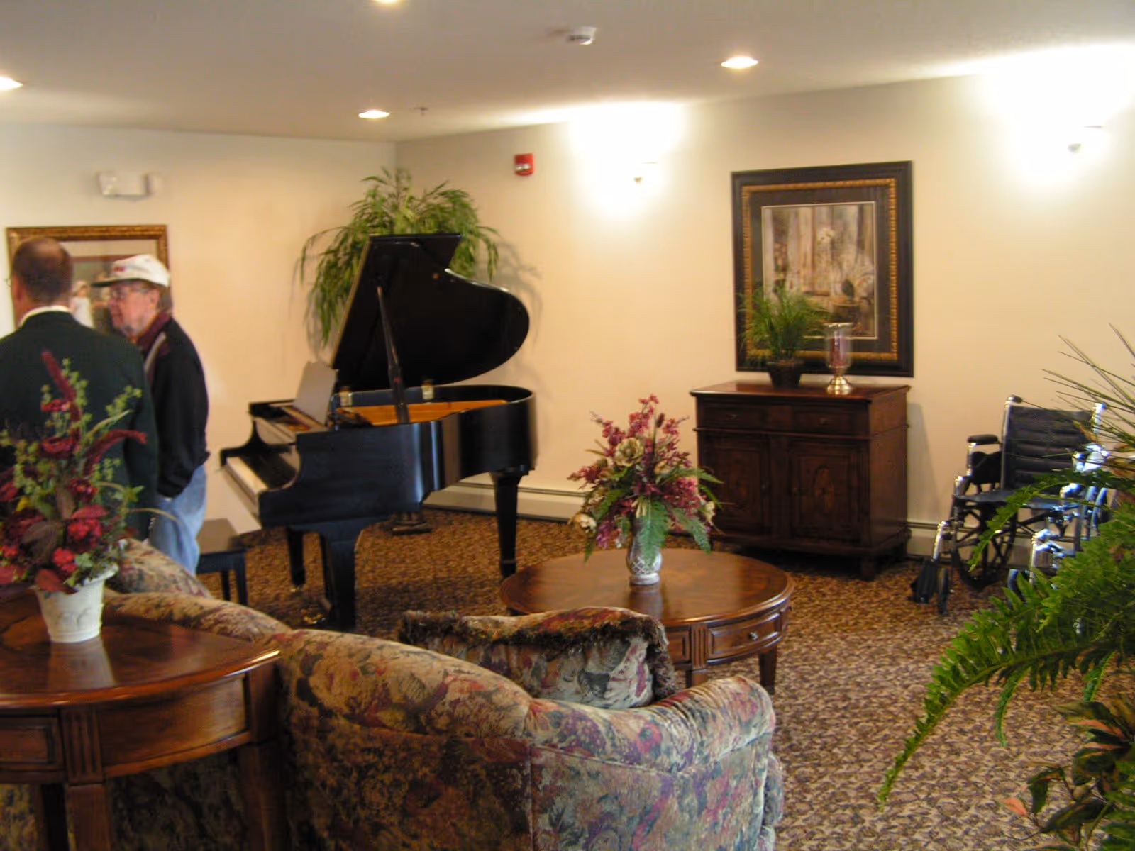 A cozy living room area in an assisted living community featuring a grand piano, floral-patterned sofas, wooden tables with flower arrangements, a wooden cabinet with a framed painting and plants, and two elderly men standing and conversing. A wheelchair is visible near the cabinet.