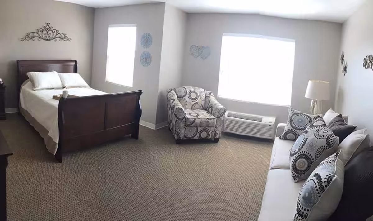 A senior living facility room featuring a wooden bed with white bedding, a patterned armchair, a white sofa with multiple decorative pillows, a lamp on a side table, and two large windows letting in natural light. The walls are decorated with metal wall art.