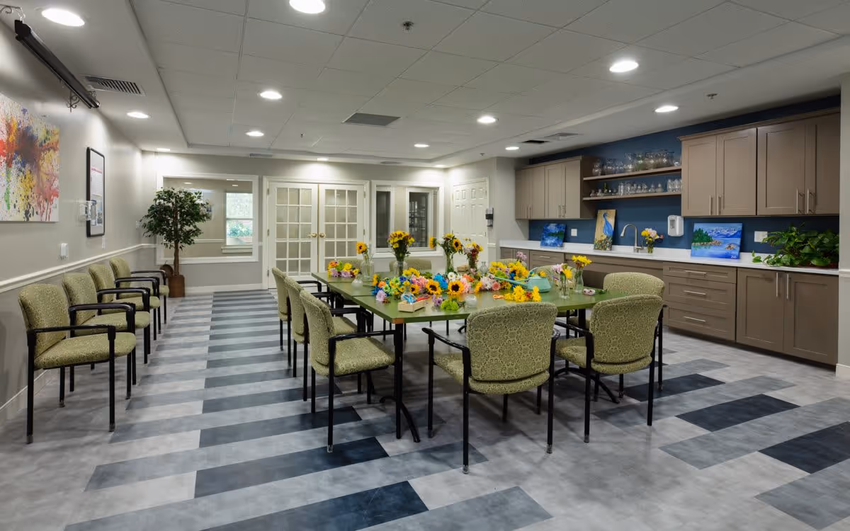 A spacious, well-lit activity room with a large table in the center surrounded by green cushioned chairs. The table is decorated with colorful artificial flowers and craft supplies. Along one wall, there are cabinets, shelves with glassware, and paintings. The floor has a striped pattern in shades of gray and blue. Additional chairs line the opposite wall, and a potted plant is placed near a window and double glass doors.