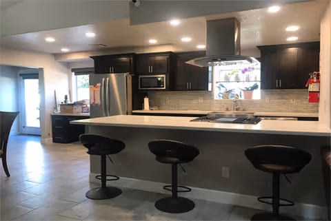 Modern kitchen with a large white island countertop featuring three black bar stools. The kitchen has dark wooden cabinets, a stainless steel refrigerator, a built-in microwave, and a stove with a stainless steel range hood. There is a window above the sink with potted plants on the windowsill, and recessed lighting in the ceiling.