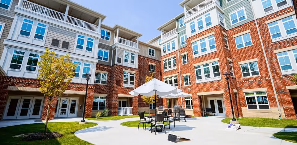 Outdoor courtyard area of a senior living facility with brick and white-paneled multi-story buildings surrounding a paved patio. The patio has tables with umbrellas and chairs, small trees, and green grass patches under a clear blue sky.
