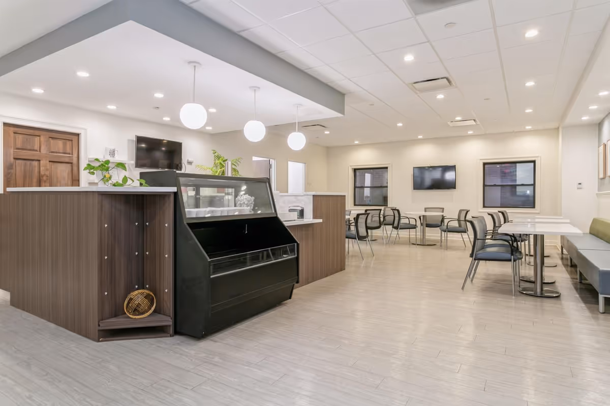 A bright and modern dining area in a senior living community featuring multiple tables with chairs, a long bench along one wall, two wall-mounted televisions, and a counter with a display case and plants. The room has light-colored flooring and ceiling with recessed lighting and hanging globe pendant lights.