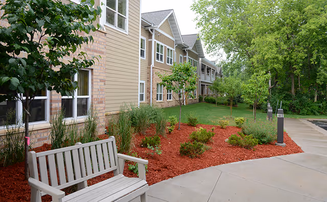 Outdoor garden area beside a senior living facility building with a beige and brick exterior. The garden features small trees, shrubs, and red mulch, with a light gray wooden bench on a concrete pathway. The area is surrounded by green grass and trees in the background.