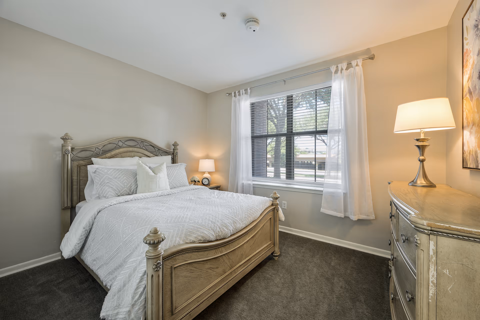 A cozy bedroom with a wooden bed frame and white bedding, a nightstand with a lamp and alarm clock, a window with white curtains letting in natural light, and a wooden dresser with a lamp and artwork above it.
