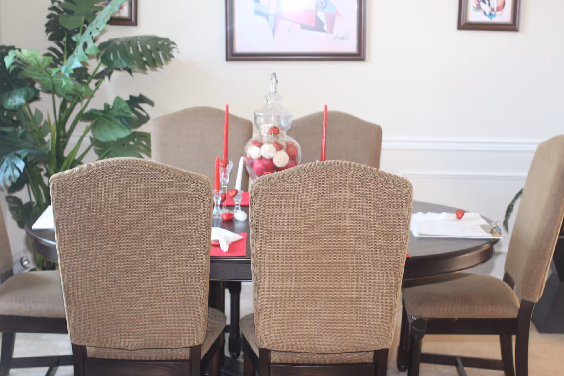A dining room with a round dark wooden table surrounded by six beige upholstered chairs. The table is decorated with red candles, a glass jar filled with decorative balls, and red placemats. There is a large green plant in the corner and framed artwork on the wall behind the table.