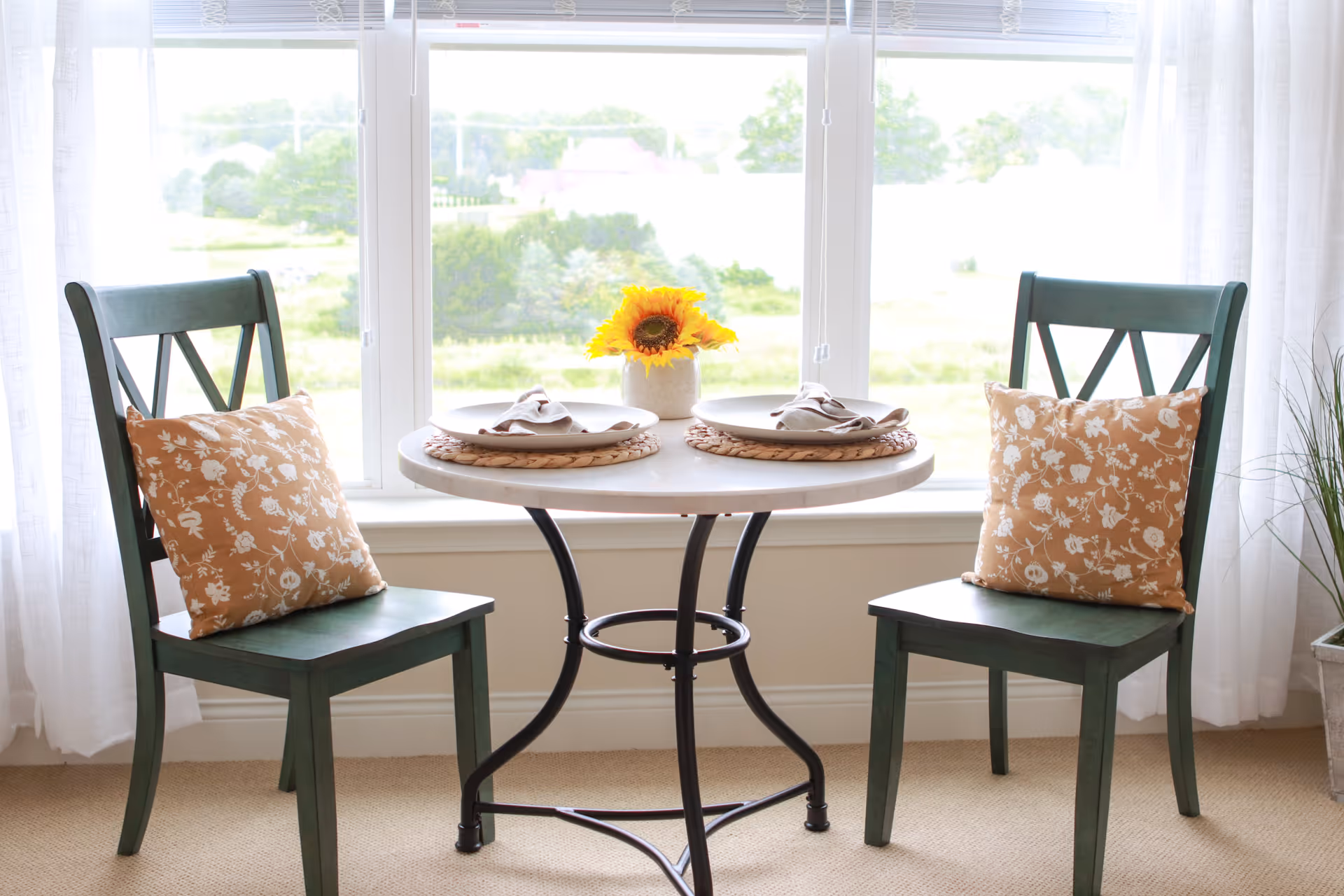A small round dining table set for two with beige placemats, white plates, and folded napkins. The table has a small vase with a sunflower in the center. Two green wooden chairs with orange floral patterned cushions are placed on either side of the table. The setting is in front of a large window with white curtains, overlooking a green outdoor landscape.