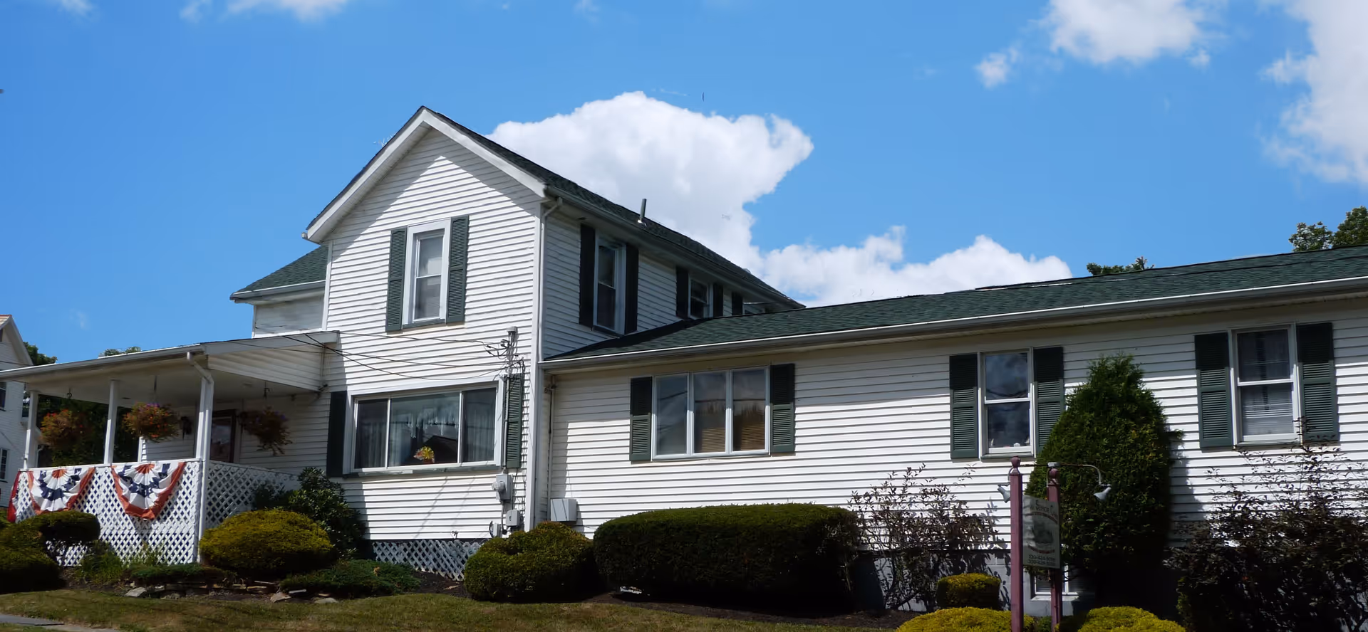 White two-story building with green shutters and a porch decorated with patriotic bunting, surrounded by shrubs under a blue sky.
