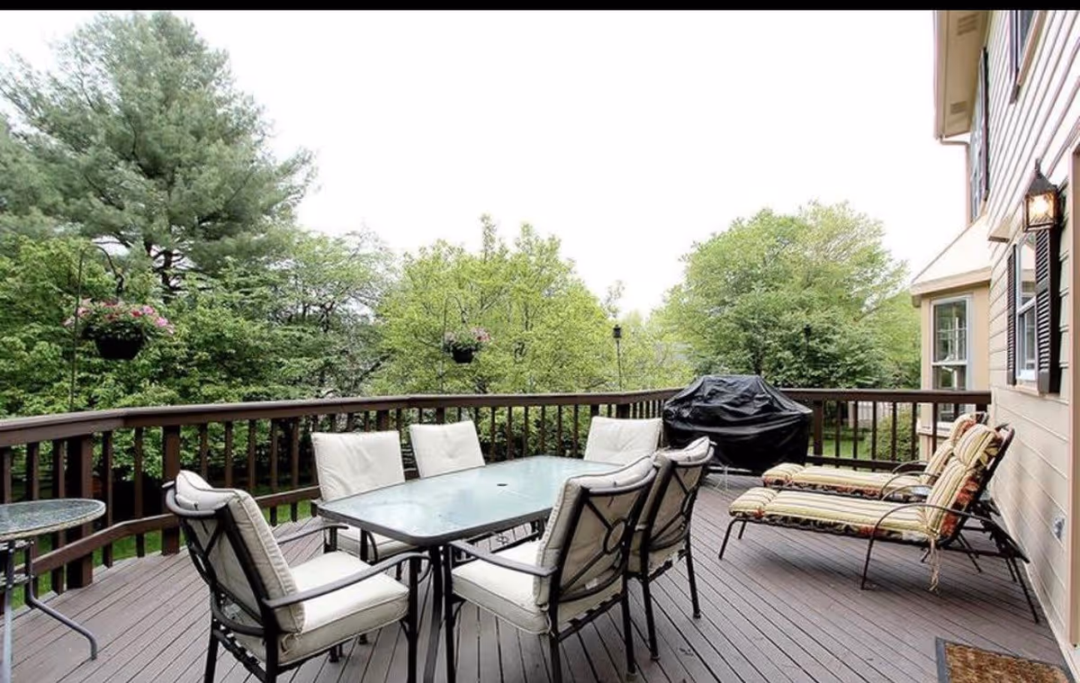 Outdoor wooden deck with a glass-top table surrounded by six cushioned chairs, two cushioned lounge chairs with striped pillows, a covered barbecue grill, and hanging flower pots. The deck overlooks a green, tree-filled yard.