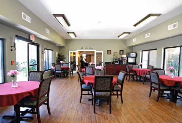 Spacious assisted-living dining room with round tables draped in red tablecloths, chairs, large windows, and wood flooring.