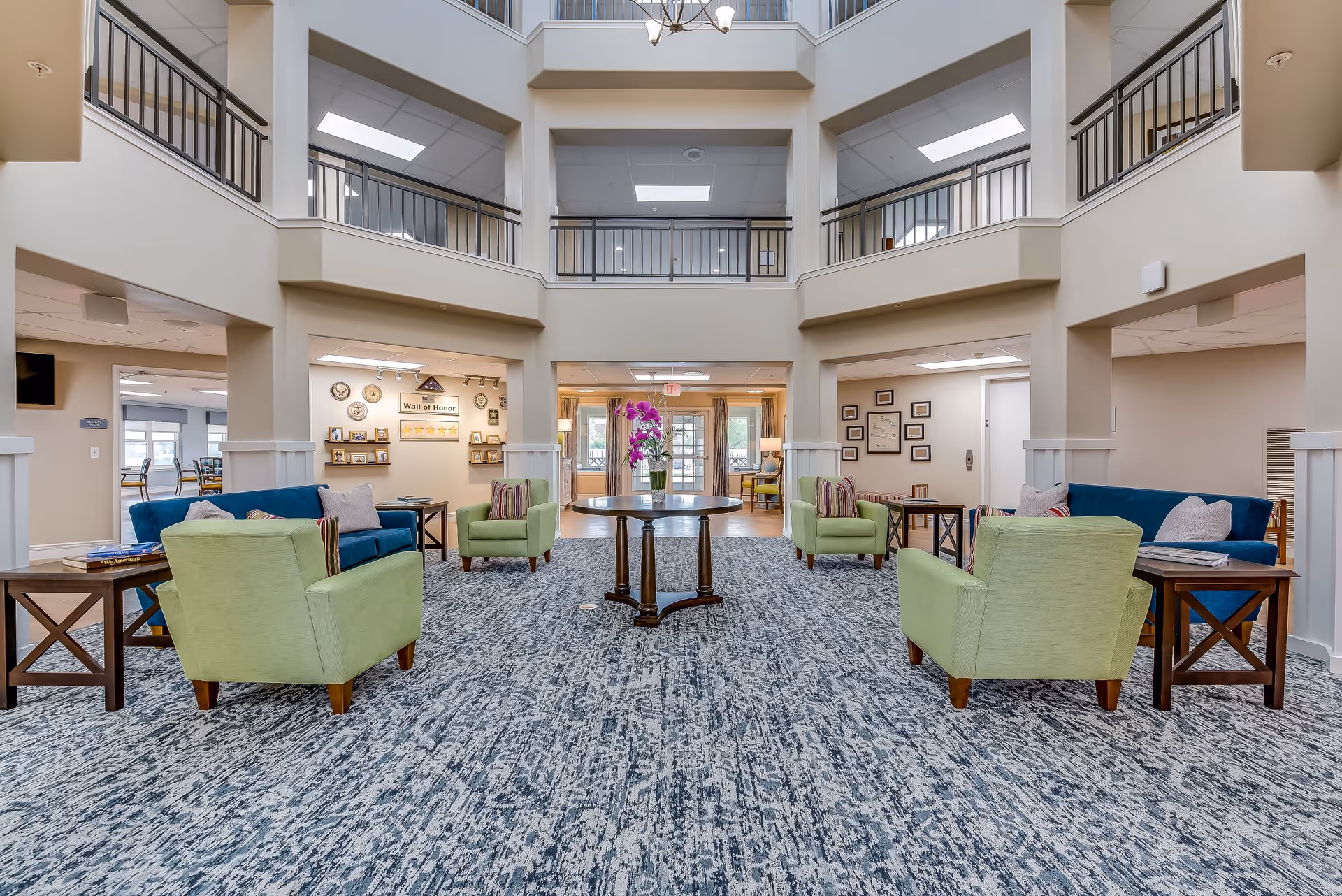 Spacious senior living facility common area with a high ceiling and balcony railings on the upper floor. The room features a patterned carpet, two blue sofas, four green armchairs arranged around a central round wooden table with a vase of purple flowers. Side tables with books and lamps are placed next to the seating. The background shows a hallway with framed pictures and a 'Wall of Honor' display.