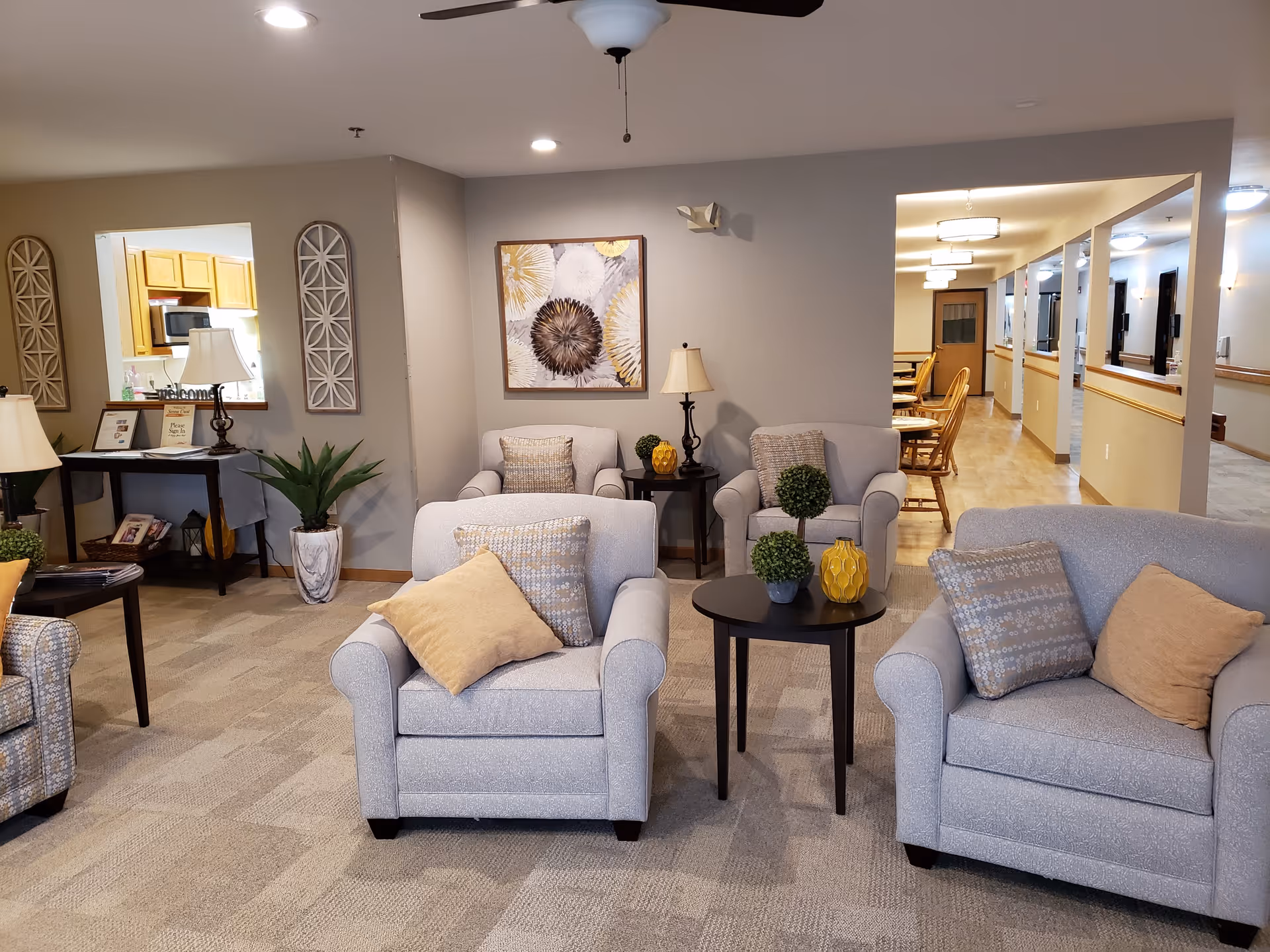 A cozy assisted living facility common area with several cushioned armchairs and small round tables decorated with small plants and yellow vases. The room has neutral-colored walls and carpeted flooring. There is a hallway leading to a dining area with wooden chairs and tables. A console table with lamps, framed pictures, and decorative items is visible near a pass-through window to the kitchen.