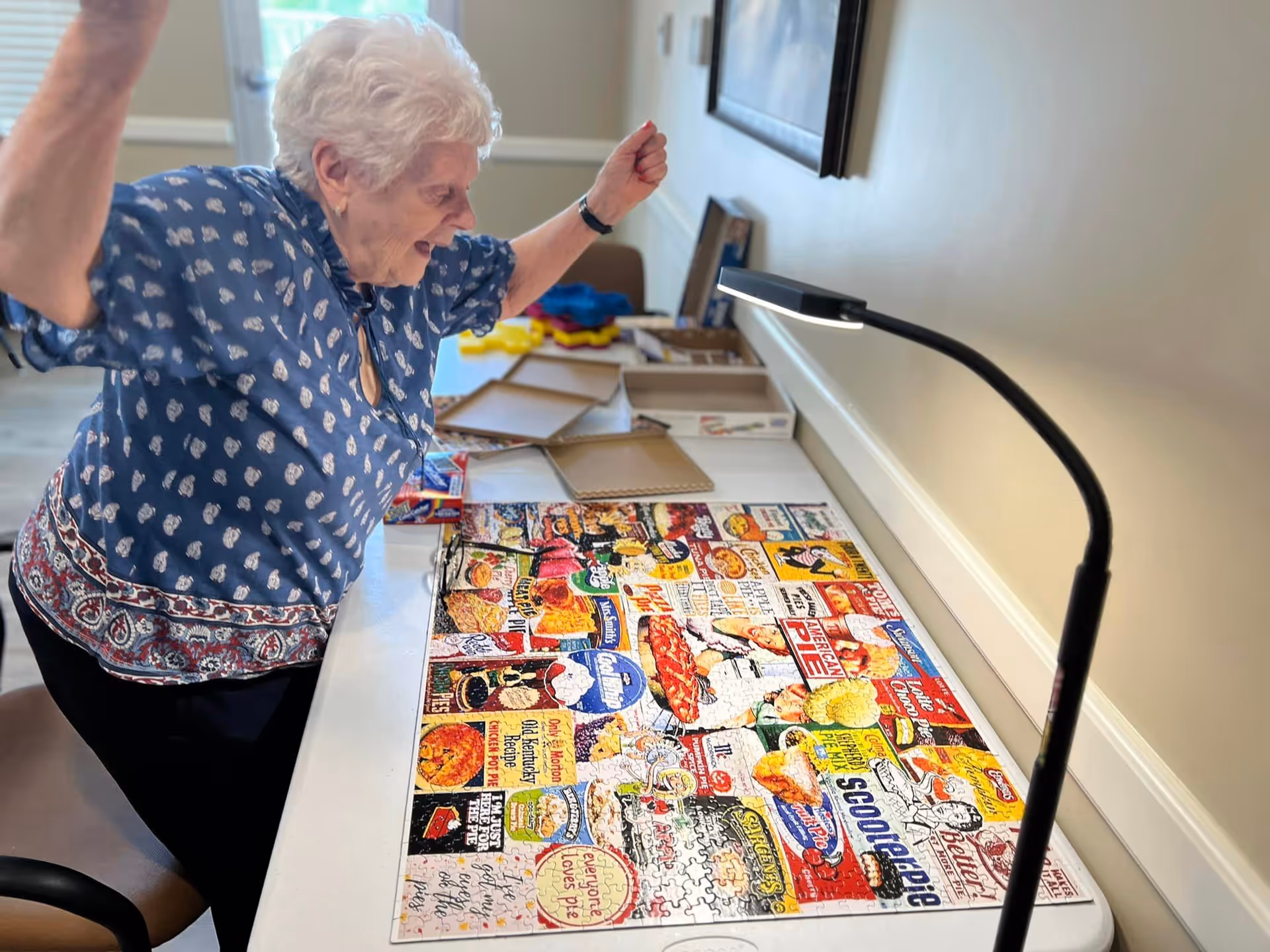 An elderly woman with white hair wearing a blue patterned blouse is joyfully raising her arms while standing next to a table with a nearly completed colorful puzzle featuring vintage food and pie advertisements. The table is in a well-lit room with a lamp focused on the puzzle.