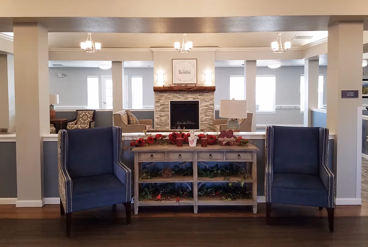 A cozy senior living room area featuring two blue upholstered armchairs with nailhead trim positioned on either side of a wooden console table decorated with red flowers and greenery. Behind the table is a stone fireplace with a wooden mantel, above which hangs a framed sign that reads 'welcome home'. The room is softly lit by ceiling chandeliers and wall sconces, with additional seating visible in the background near windows.