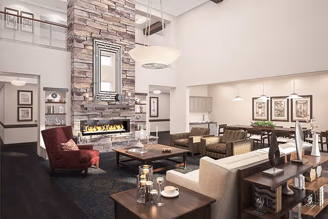 A spacious and elegant living room area with a tall stone fireplace as the focal point. The room features a high ceiling with exposed beams, a large mirror above the fireplace, and a variety of seating including a red armchair, beige sofa, and brown armchairs. There are several tables with decorative items such as candles, vases, and books. The room is well-lit with pendant lights and has framed artwork on the walls.