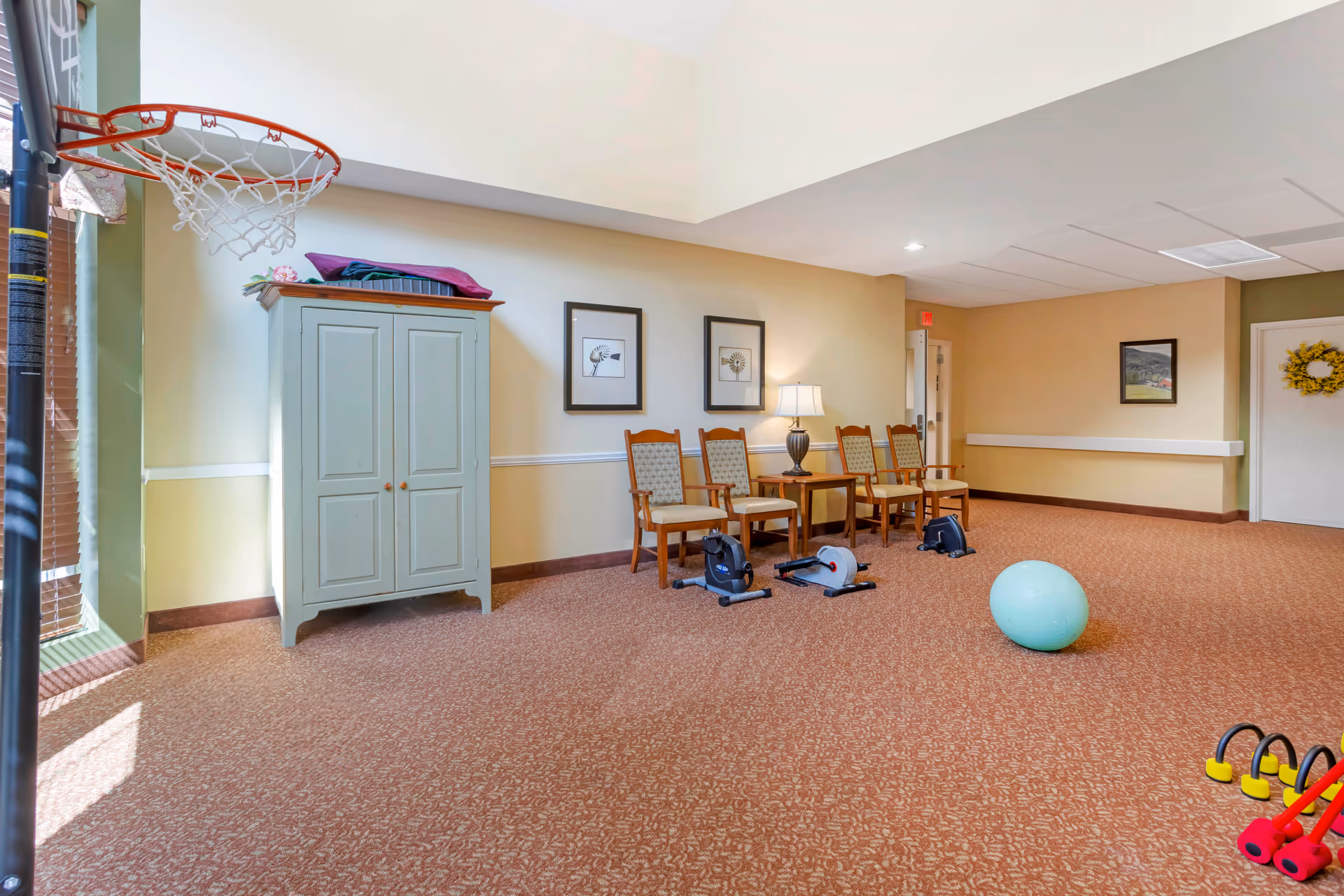 A spacious room with beige walls and carpeted floor featuring a small basketball hoop on the left, a light green cabinet, four wooden chairs with patterned cushions, a table with a lamp, exercise equipment including pedal exercisers and a large blue exercise ball, and a set of colorful dumbbells on the floor.