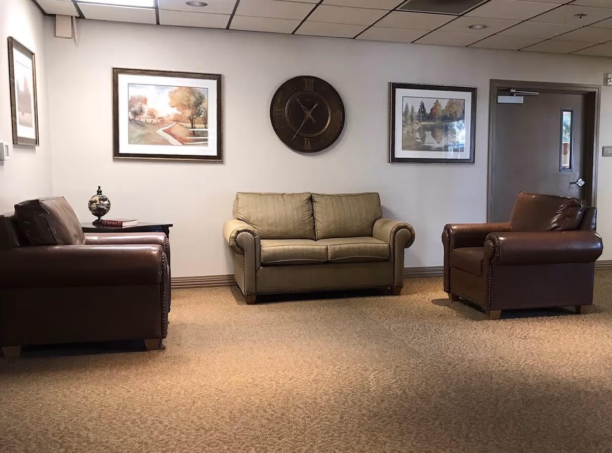 A seating area in a senior living facility with two brown leather armchairs and a beige fabric loveseat arranged on a carpeted floor. The wall behind the seating has a large round clock in the center and two framed landscape paintings on either side. There is a side table with a decorative globe and a book next to one of the armchairs. A closed door is visible on the right side of the image.