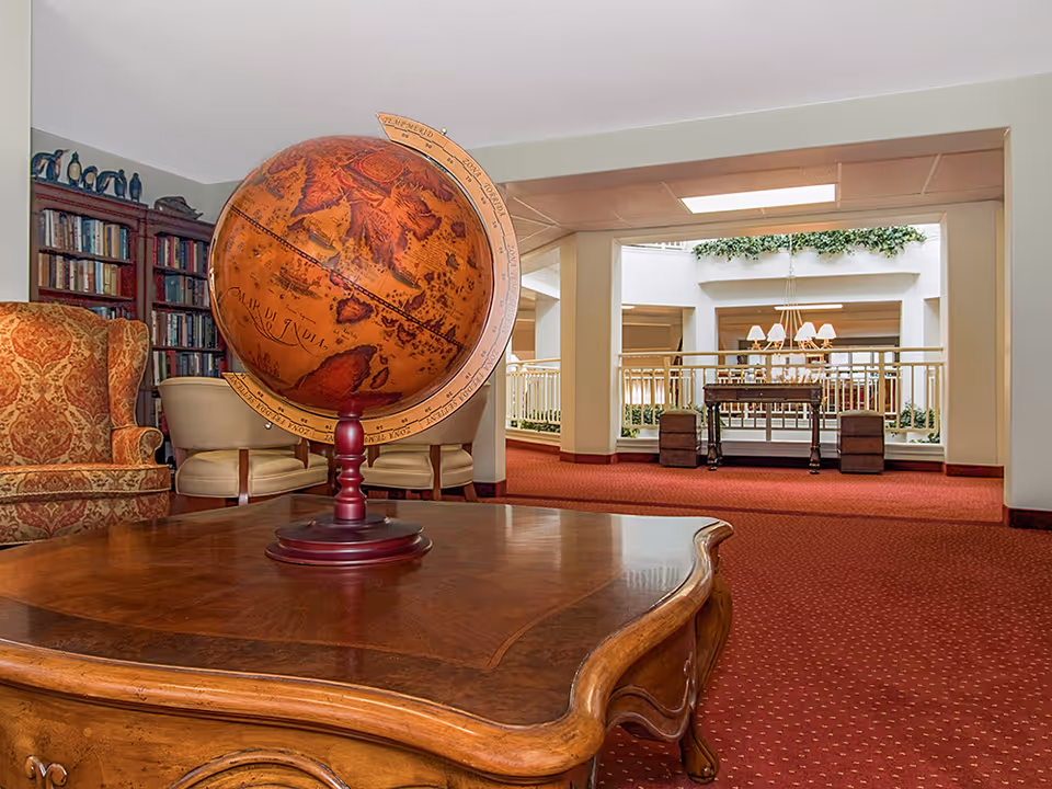 A cozy interior lounge area featuring a large vintage-style globe on a wooden coffee table. In the background, there is a patterned armchair, bookshelves filled with books, and cream-colored chairs. The space opens up to a well-lit atrium with a hanging chandelier and greenery along the upper ledge.