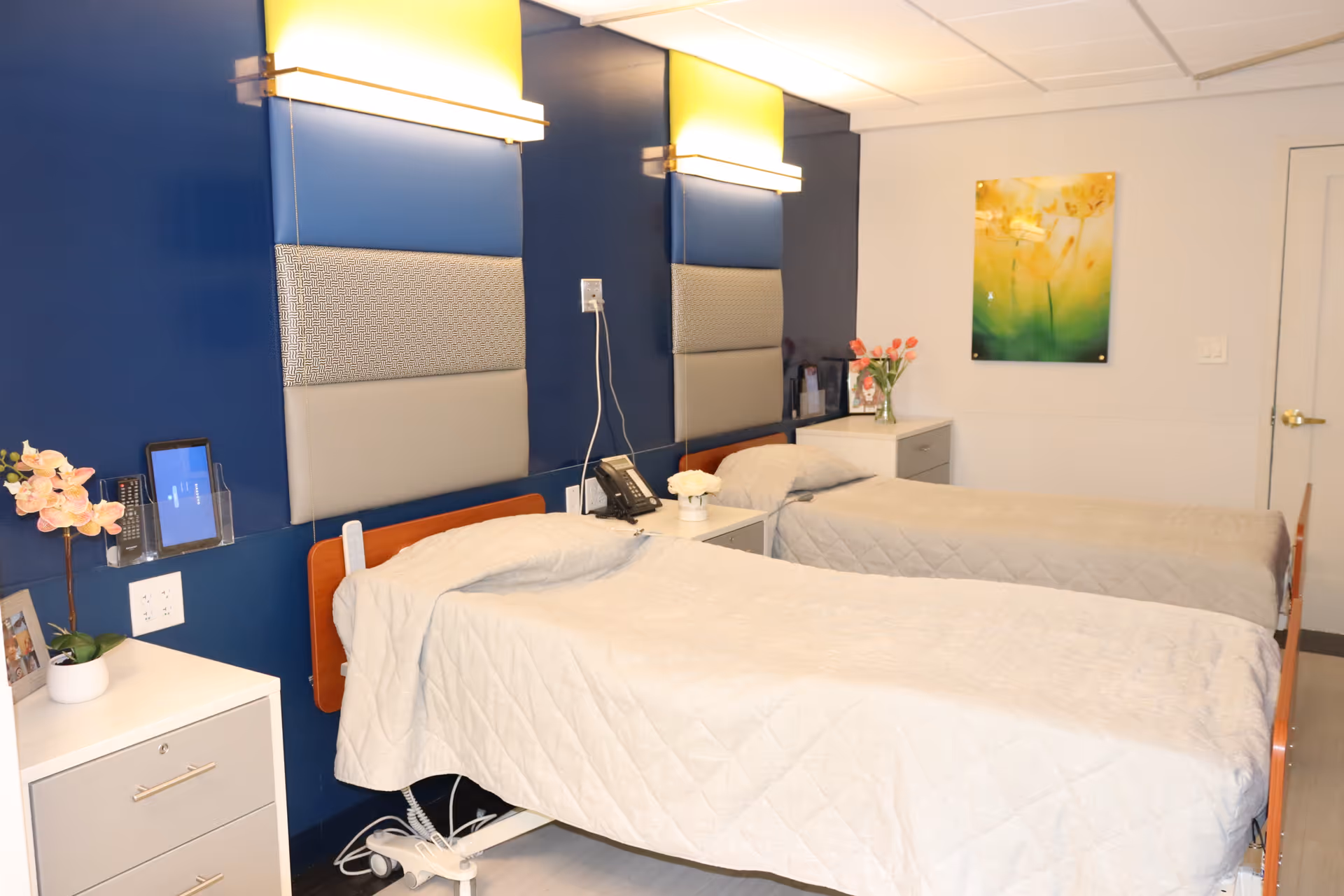A clean and well-lit room with two hospital-style beds covered in white quilts. Each bed has a padded headboard mounted on a dark blue wall with a light fixture above. There are two white bedside tables with flowers and a telephone on them. A colorful floral painting hangs on the white wall in the background.