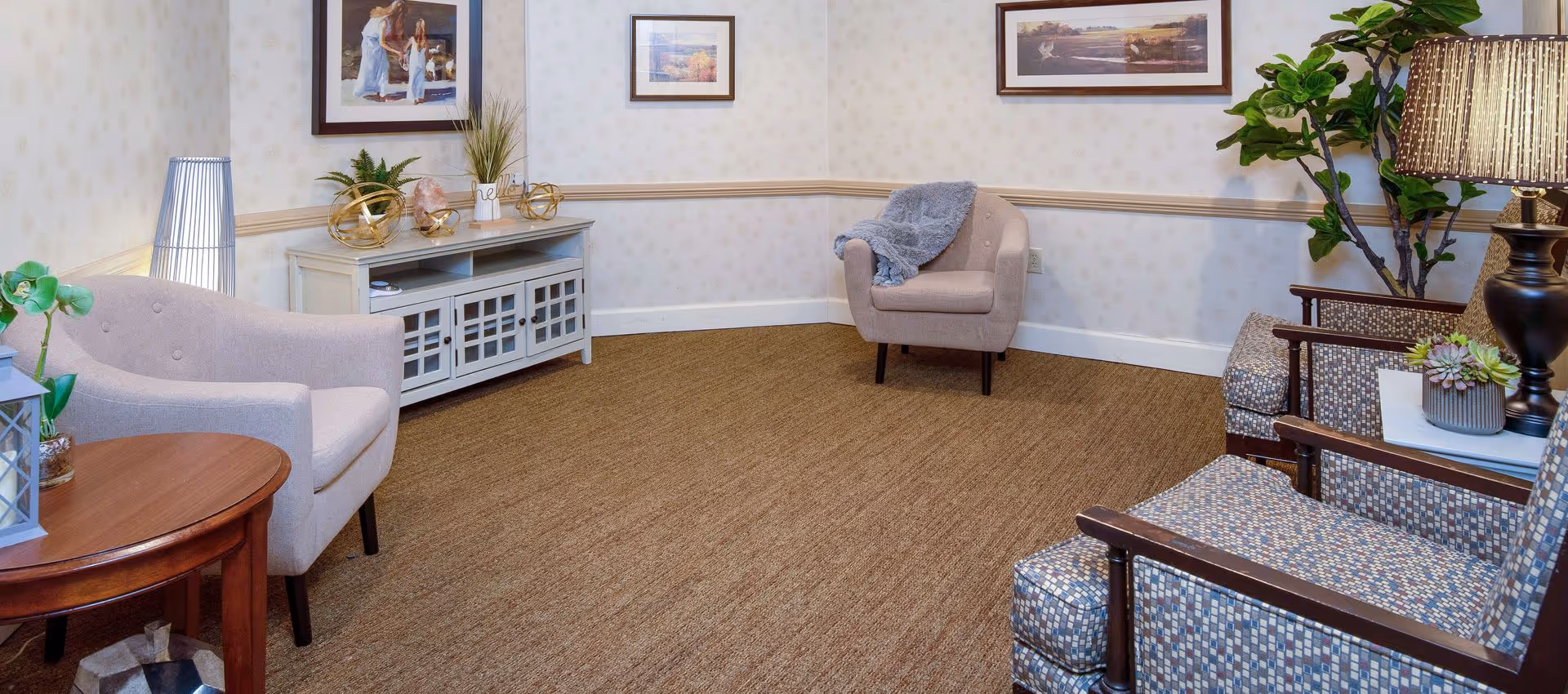 A cozy sitting area in a senior living facility with beige armchairs, a wooden side table, a white cabinet with decorative items, framed artwork on the walls, a floor lamp, and a potted plant.
