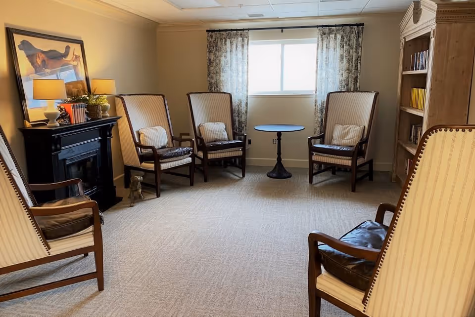 A cozy sitting room with six high-back armchairs arranged in a semi-circle around a small round table near a window with patterned curtains. There is a black fireplace mantel with two lamps and decorative items, a framed picture above it, and a wooden bookshelf filled with books on the right side of the room. The room has beige walls and carpeted flooring.