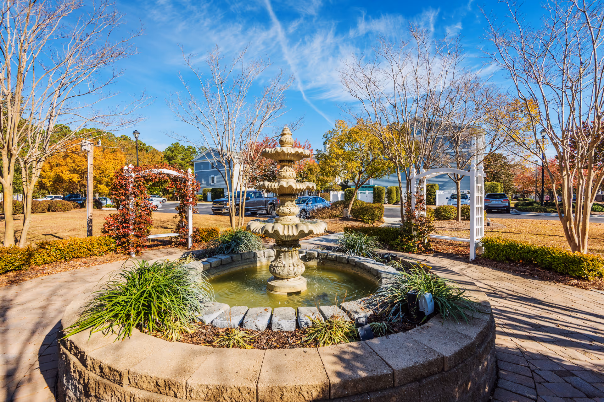 Outdoor garden area with a multi-tiered stone fountain in the center, surrounded by a circular stone border and greenery. There are leafless trees and bushes with autumn foliage, a paved walkway, and white wooden trellises with climbing plants. Several parked cars and residential buildings are visible in the background under a blue sky with wispy clouds.