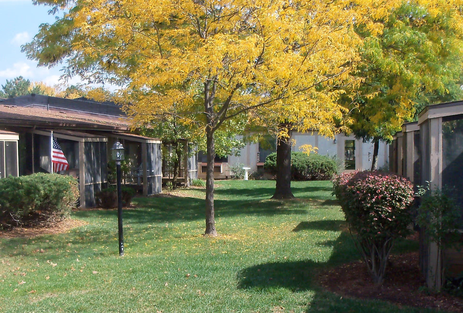 A grassy courtyard area with a tree that has yellow autumn leaves in the center. There are bushes and shrubs along the sides, screened porch structures on both sides, and an American flag visible on the left side. The sky is partly cloudy.