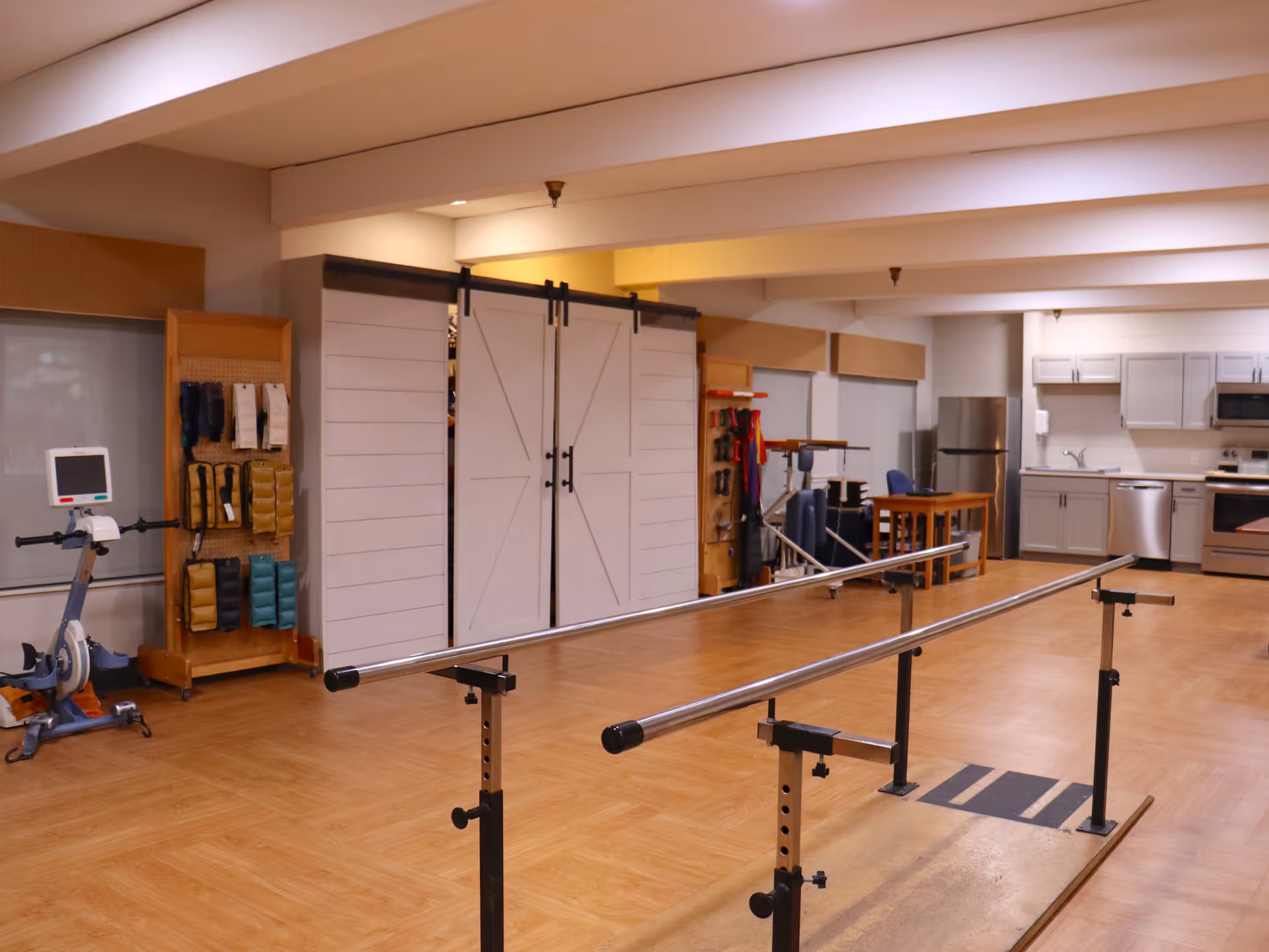 Rehabilitation therapy room with parallel walking bars, exercise equipment, and a kitchenette in the background.