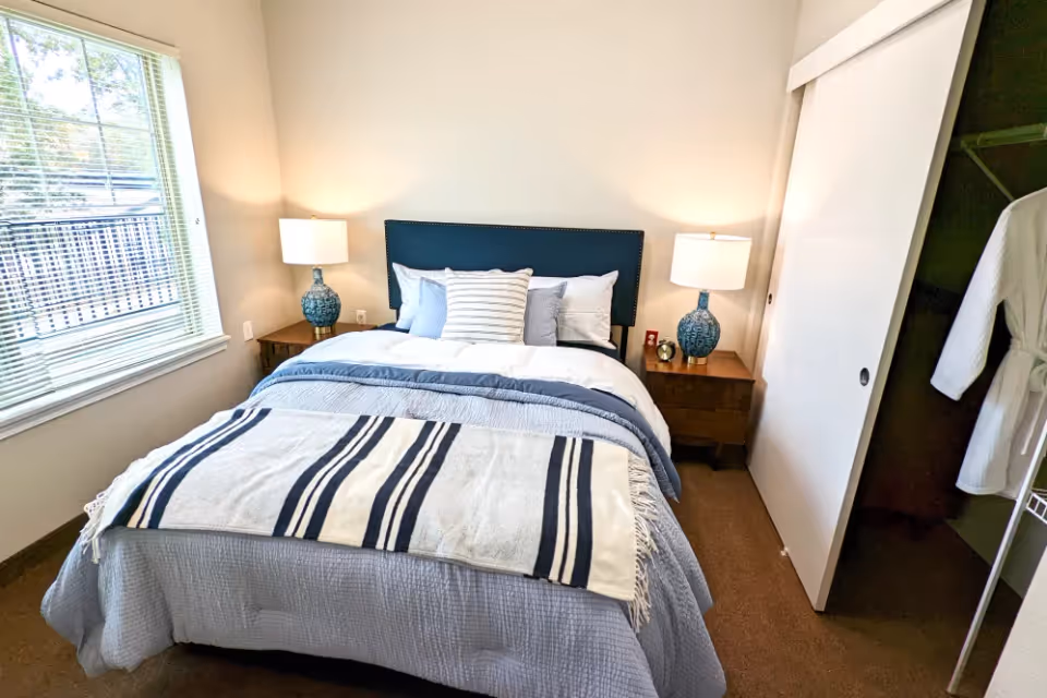 Well-lit bedroom with a centered bed dressed in blue and white linens, two bedside tables with lamps, a window to the left and an open closet to the right.