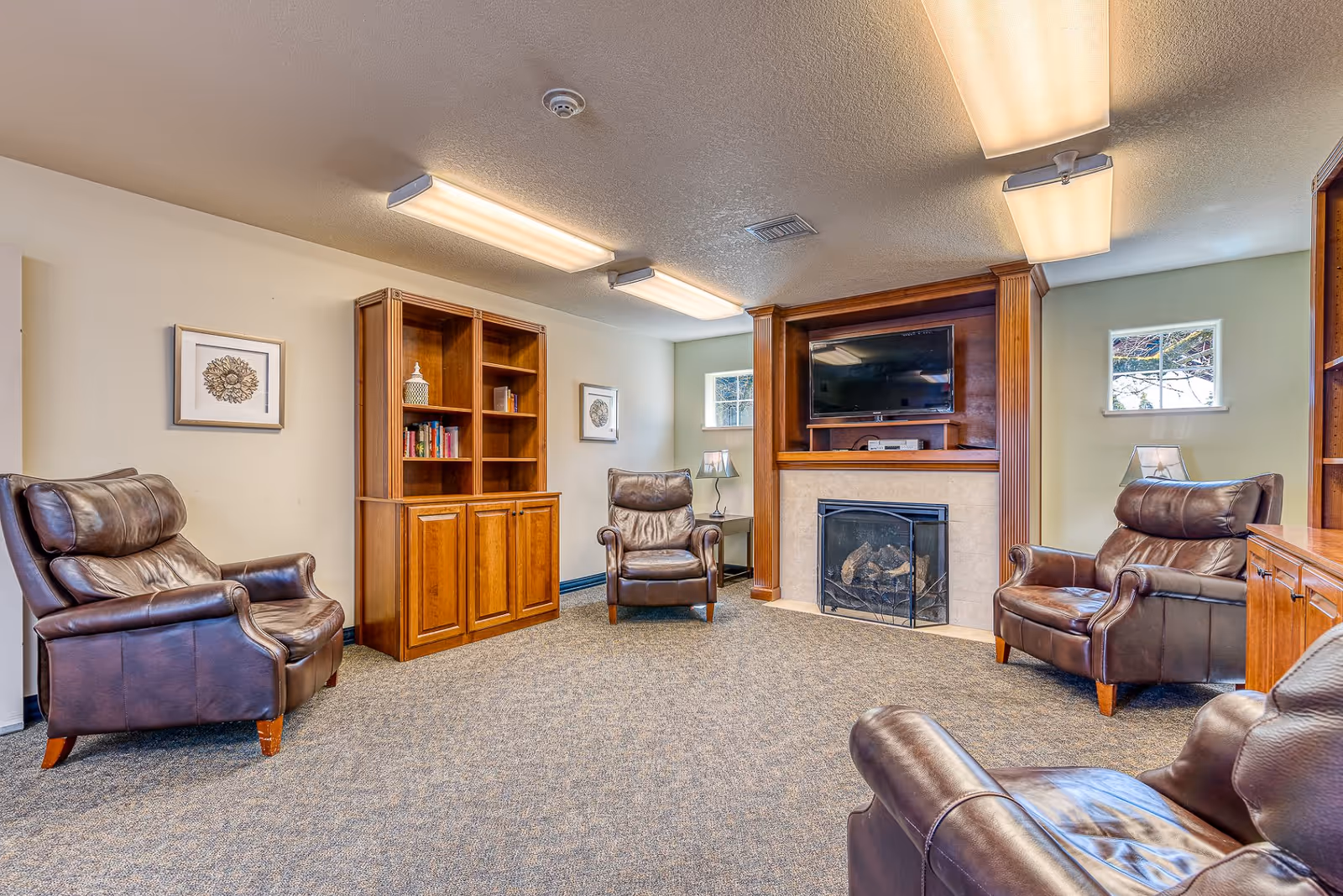 A cozy living room with four brown leather armchairs arranged around a fireplace with a TV mounted above it. The room has beige walls, carpeted floor, wooden built-in shelves with books and decorative items, two small windows, and ceiling lights.