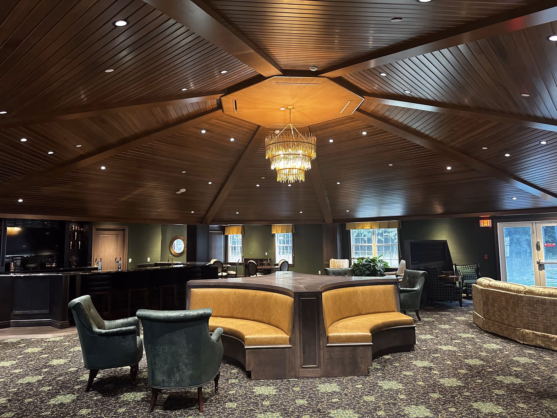 Interior view of a senior living facility lounge area with a large wooden octagonal ceiling and a chandelier in the center. The room features a curved yellow leather bench seating arrangement in the middle, green upholstered armchairs, a patterned carpet, and a bar area with stools in the background. Large windows allow natural light into the space.